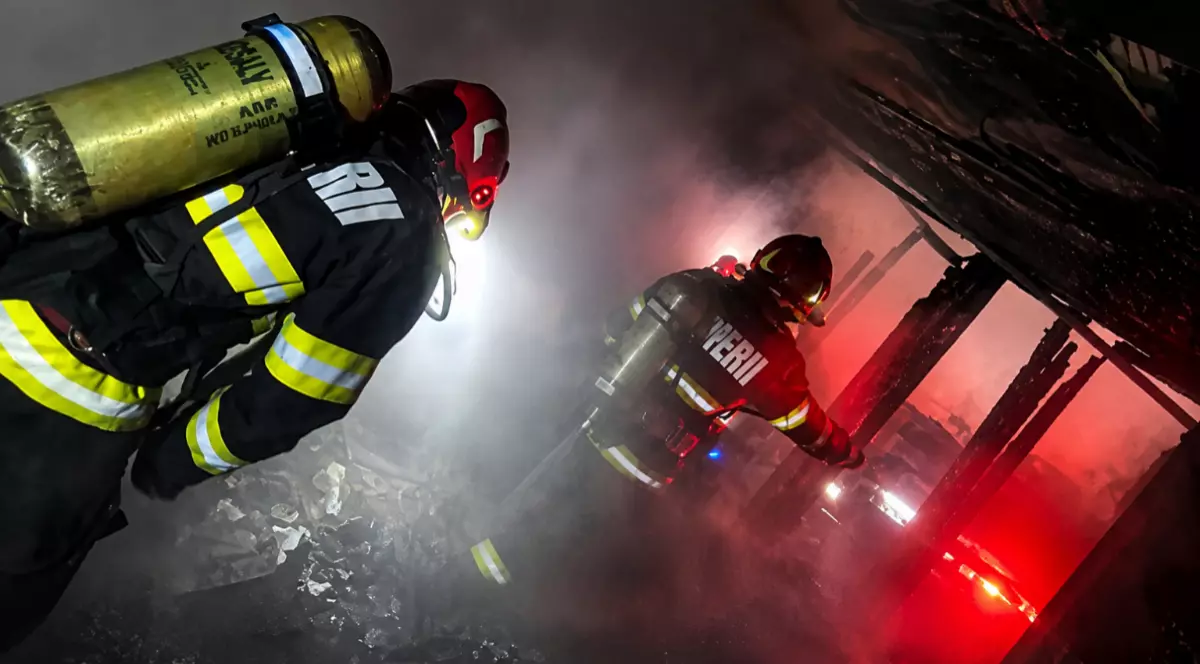 Imaginea surprinde pompieri în costume de protecție și tuburi de oxigen care intervin într-un spațiu plin de fum și flăcări pentru stingerea unui incendiu.