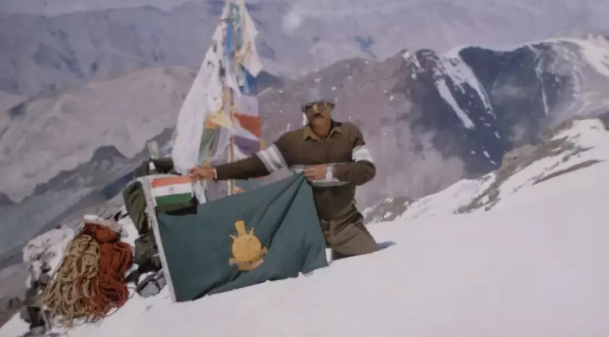 Imaginea prezintă un militar indian aflat într-o zonă montană acoperită de zăpadă, lângă steagurile Indiei și ale unei unități militare. În fundal se văd vârfuri muntoase înalte, iar în jurul său sunt echipamente de alpinism și corzi.