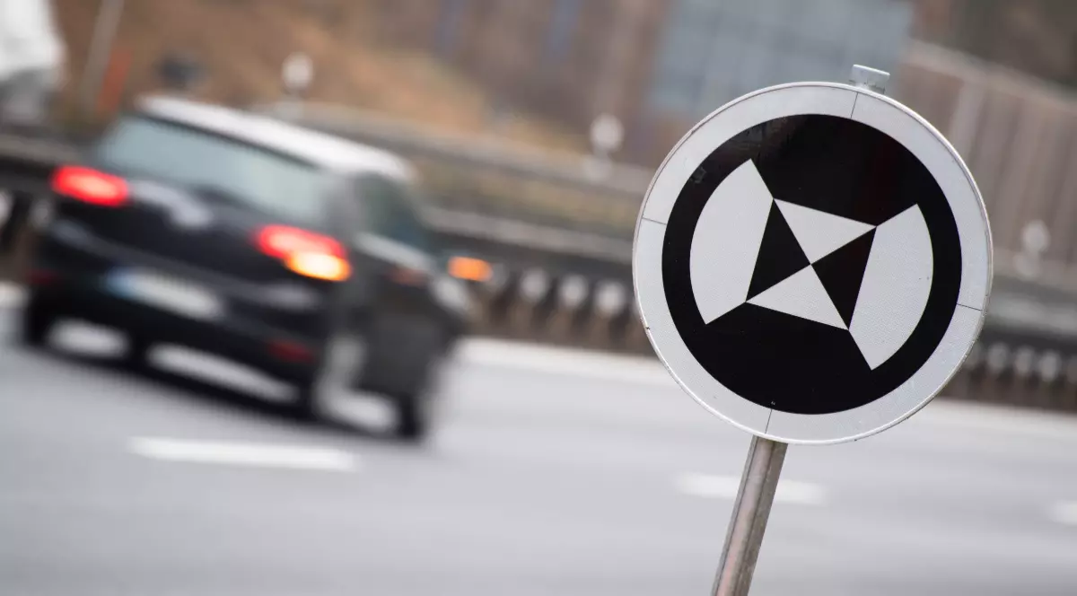 Imaginea prezintă un indicator rutier circular cu un simbol alb-negru special pentru vehicule autonome, pe fundalul estompat al unei mașini în mișcare pe o autostradă.