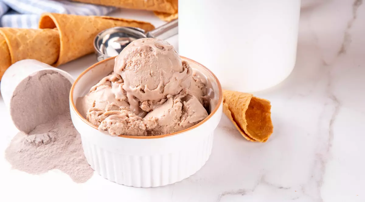 Imaginea prezintă un bol cu mai multe cupe de înghețată cu aromă de ciocolată, alături de cornete crocante și pudră de cacao. Desertul este așezat pe o suprafață deschisă la culoare, într-o compoziție luminoasă și apetisantă.