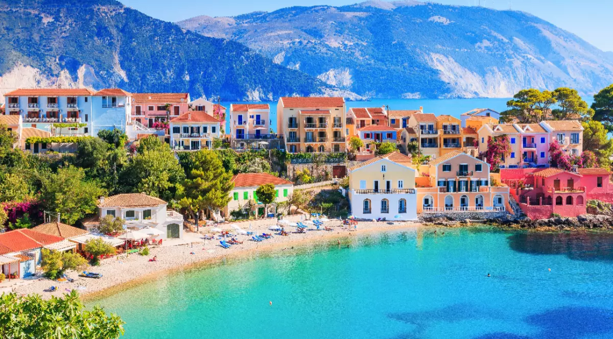 Imaginea prezintă un sat de coastă cu clădiri viu colorate, situat pe malul unui golf cu ape turcoaz și plajă, având în fundal muntele.