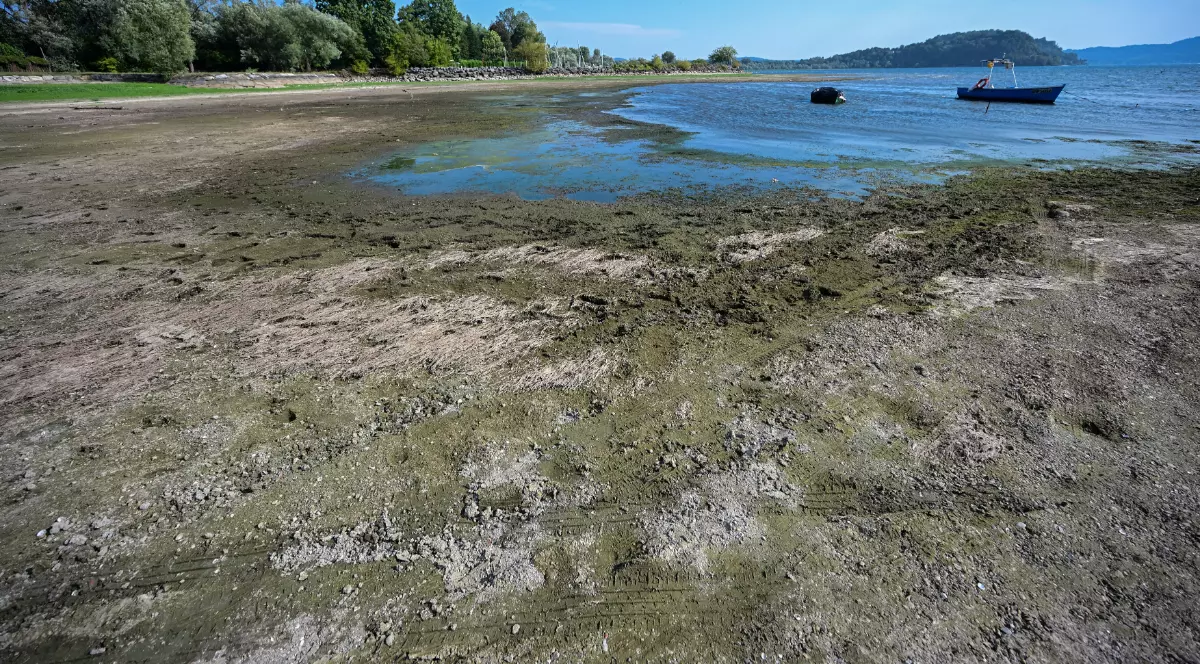 Lacul Maggiore apare cu nivelul apei mult scăzut, lăsând la vedere o porțiune întinsă de mal și nămol. În depărtare, o barcă plutește pe apa rămasă, în timp ce peisajul este dominat de vegetație și dealuri.