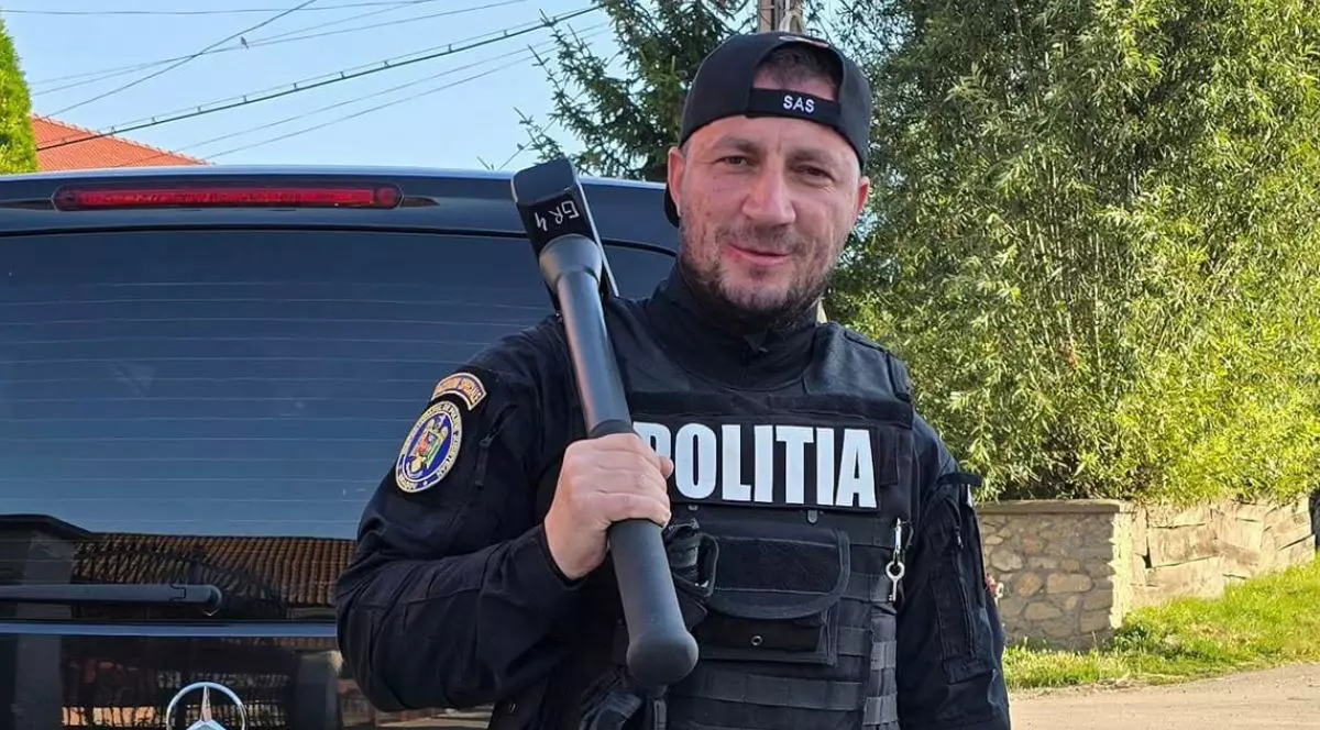 Marian Godină, într-o uniformă neagră de Poliție, cu șapca întoarsă la spate pe care scrie SAS și cu un dispozitiv de deschis uși într-o mână, pe umăr