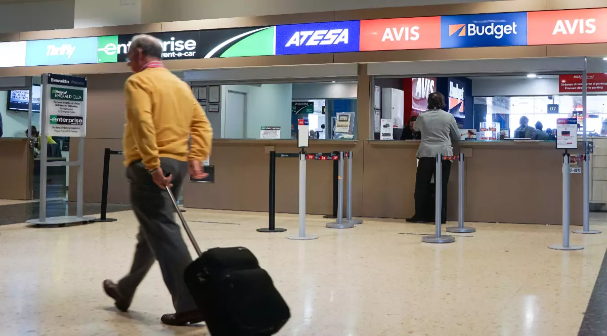 Imaginea arată ghiseele mai multor companii de închirieri auto dintr-un aeroport, în timp ce un bărbat își trage trollerul trecând prin fața lor.