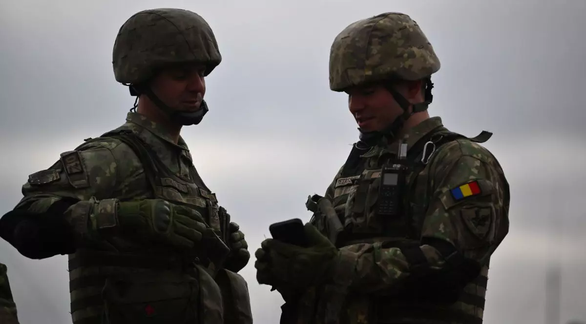Doi militari români, îmbrăcați în uniforme, cu căști pe cap, cu cerul înnorat pe fundal