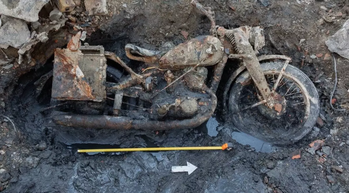 Imaginea prezintă o motocicletă veche și puternic ruginită, lângă care sunt plasate o ruletă și o săgeată indicator pentru măsurători.