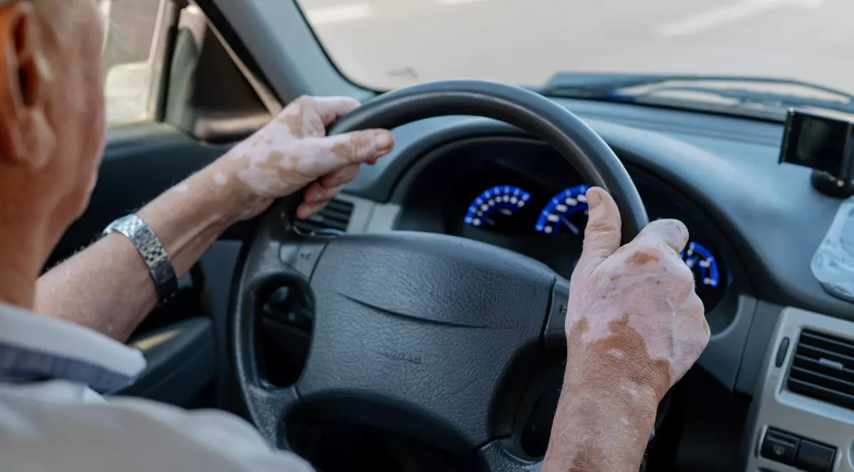 În imagine se vede o persoană în vârstă aflată la volanul unei mașini. Mâinile sale sunt poziționate pe volan, iar în față se observă bordul și drumul pe care circulă.