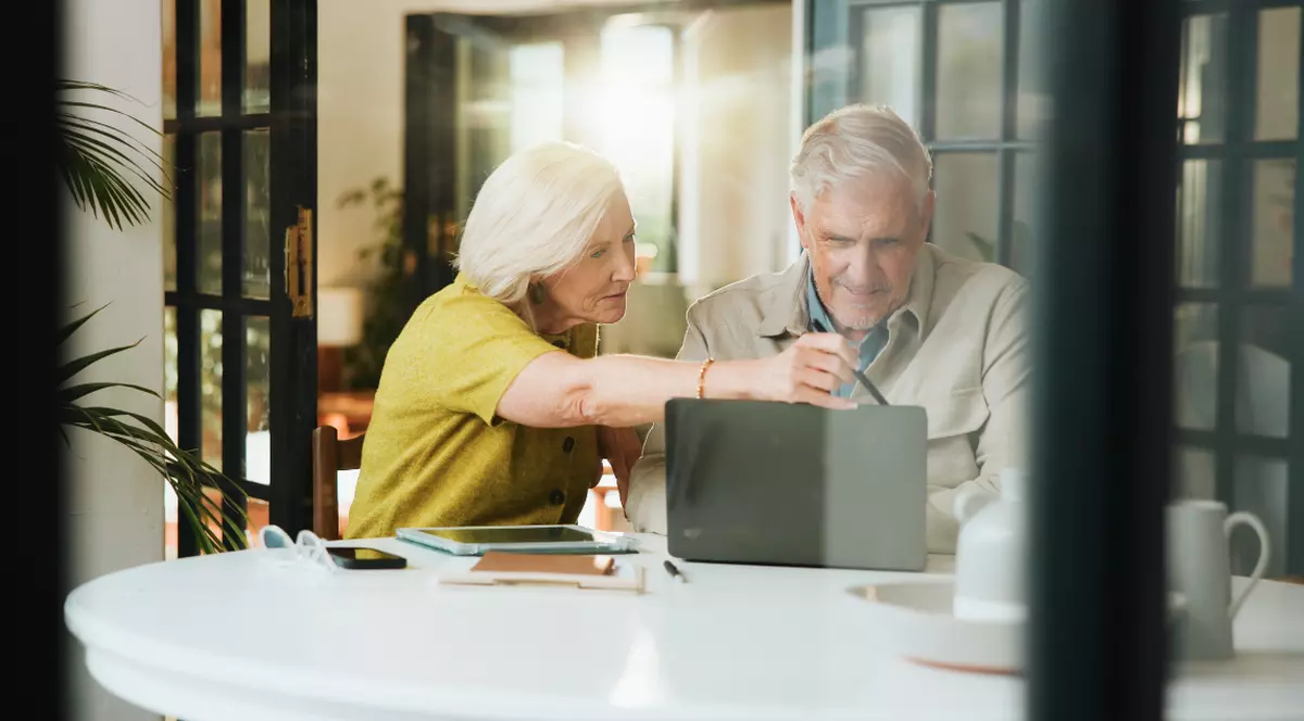 În imagine apare un cuplu de pensionari așezat la masa din bucătărie, analizând împreună informații pe un laptop și documente. Atmosfera sugerează că cei doi își gestionează cu atenție bugetul sau își planifică finanțele, colaborând într-un mod calm și organizat.