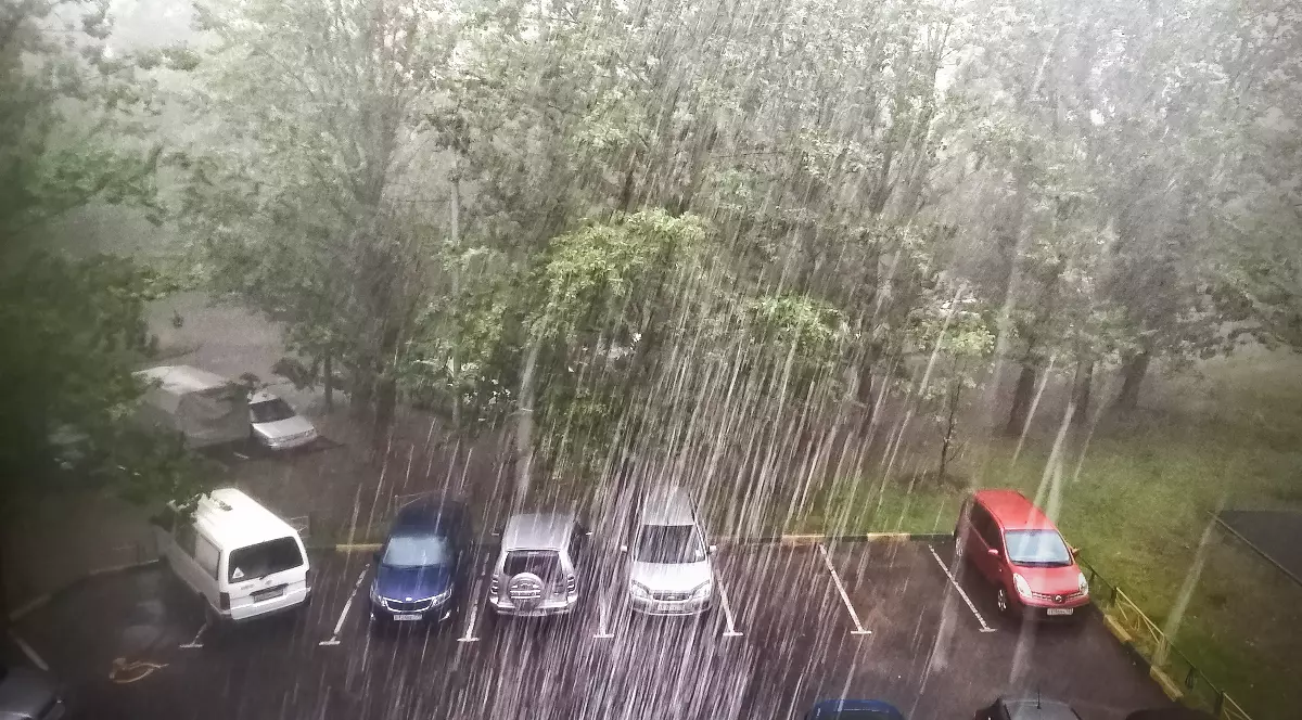 Imaginea prezintă o ploaie torențială densă căzând peste mașini într-o parcare mărginită de copaci.