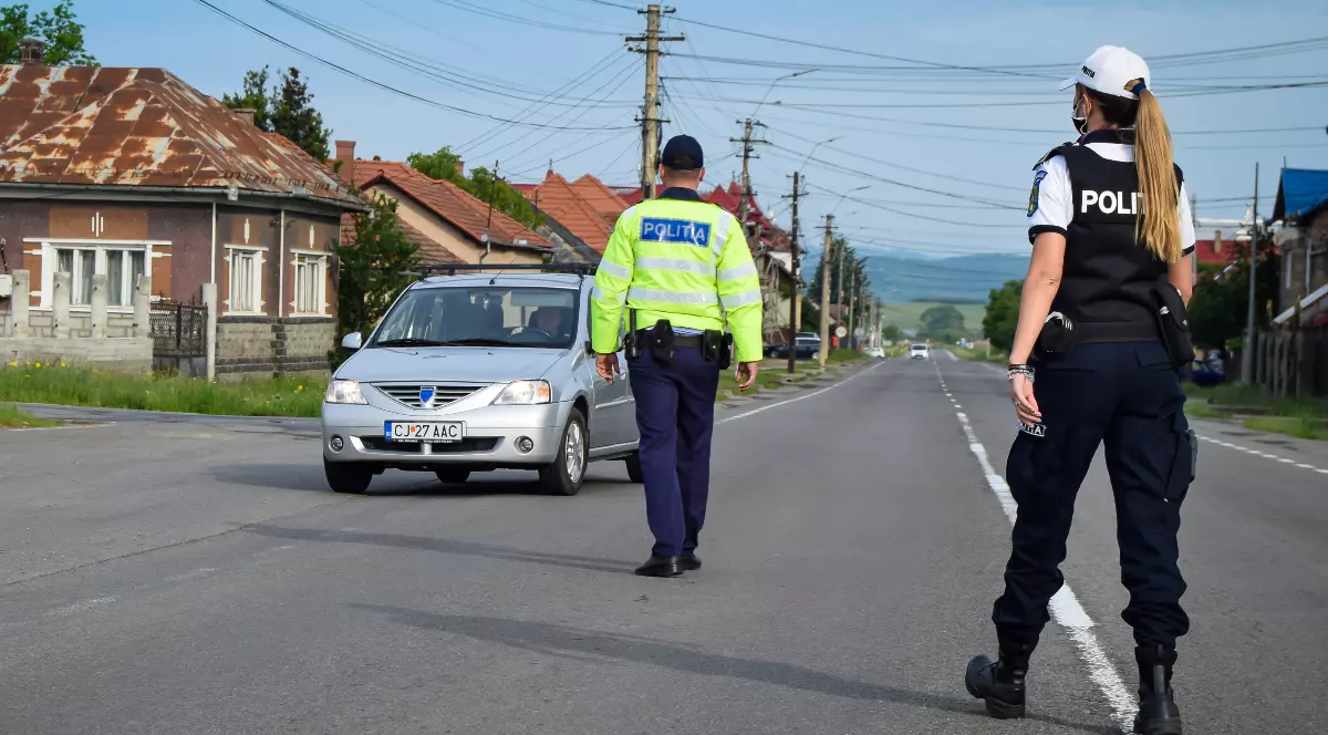 Imaginea prezintă doi polițiști români aflați pe carosabil într-o zonă rurală, dintre care unul oprește pentru control o mașină argintie.