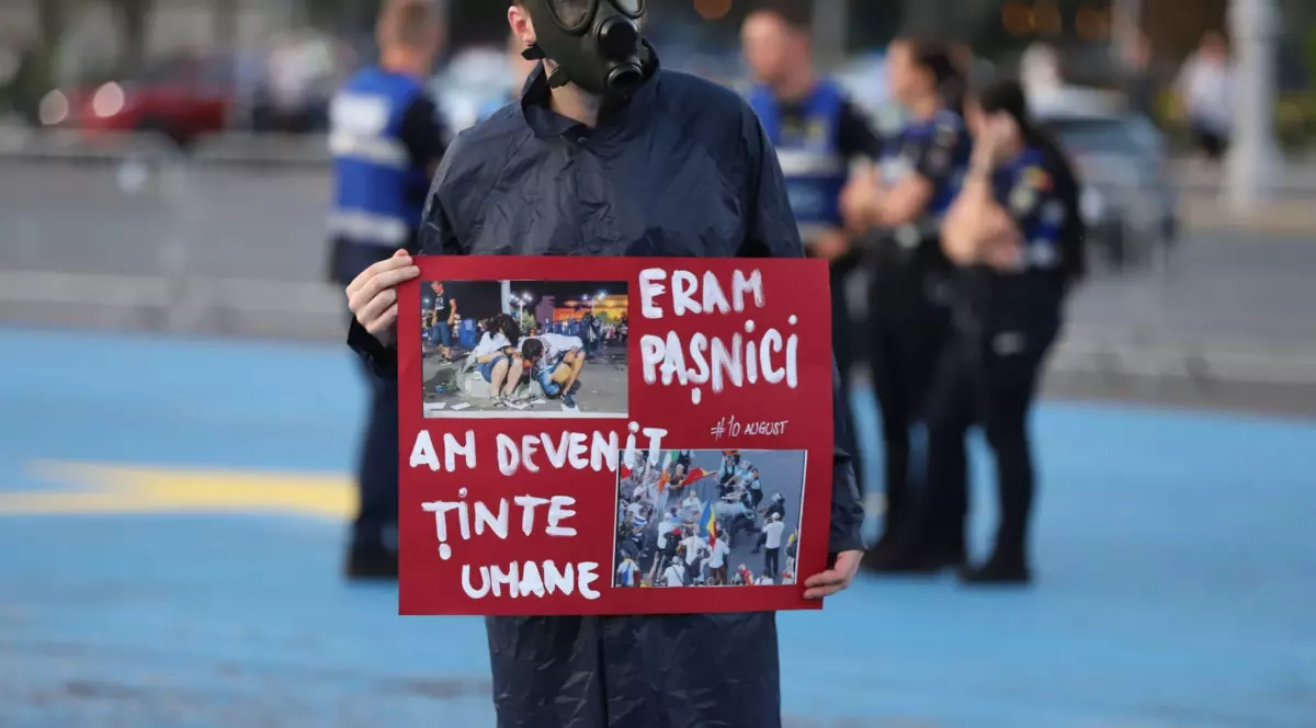 În imagine se vede un protestatar purtând o mască de gaze și ținând o pancartă roșie cu mesajul „ERAM PAȘNICI / AM DEVENIT ȚINTE UMANE”, alături de fotografii de la protestele din 10 august. În fundal se află mai multe persoane, iar scena pare să fie surprinsă în Piața Victoriei, în timpul manifestației comemorative.