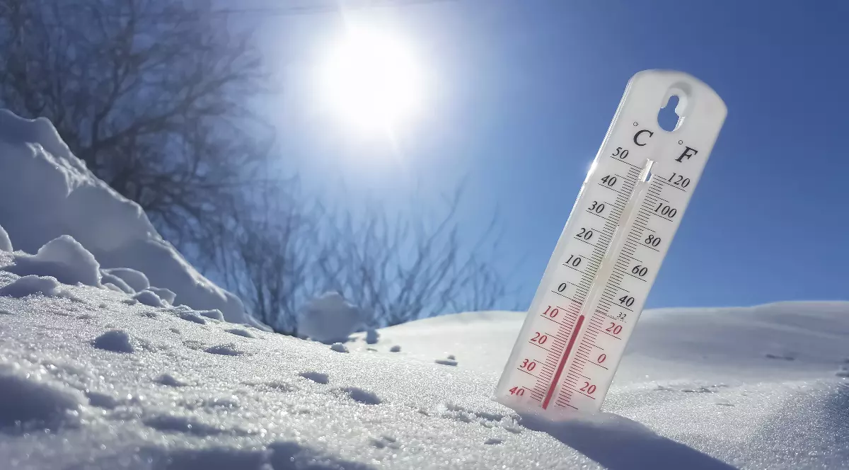 Imaginea prezintă un termometru alb înfipt în zăpadă sub un cer senin și însorit, indicând o temperatură scăzută.