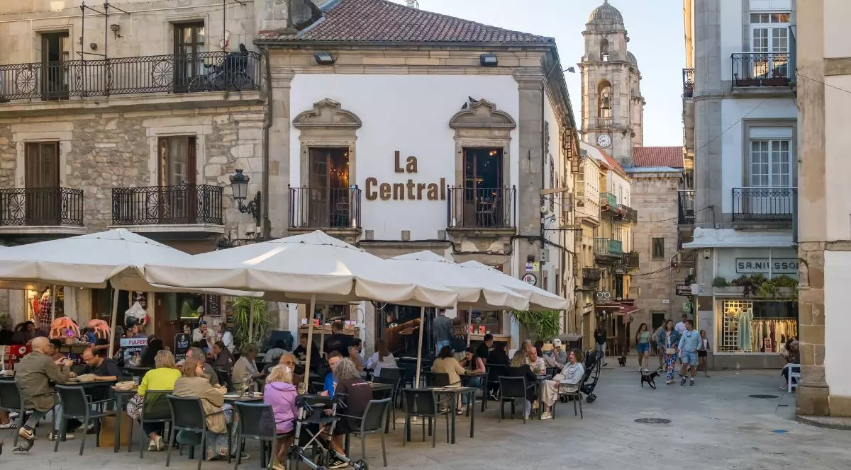 Vigo, Spania – 7 iulie 2024: O scenă plină de viață într-o piață istorică din oraș, unde oamenii stau sub umbrele albe pe terasa unui restaurant, cu turnul unei biserici în fundal.