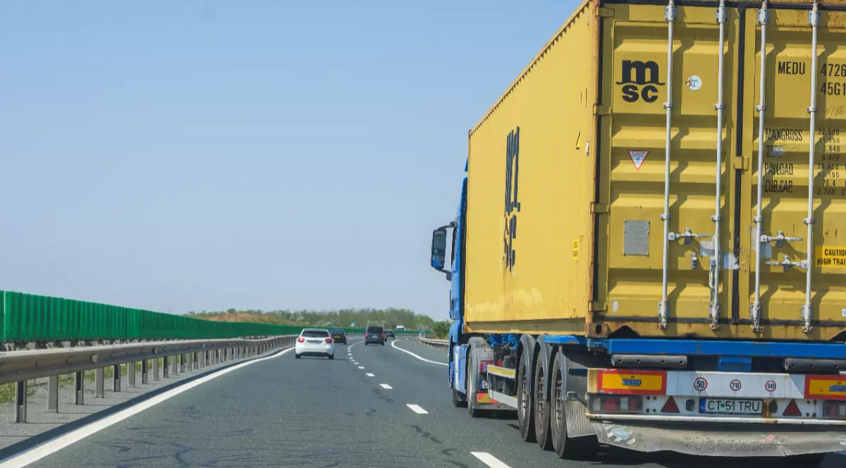 Imaginea prezintă un autotren albastru care transportă un container galben, deplasându-se pe banda din dreapta a unei autostrăzi.