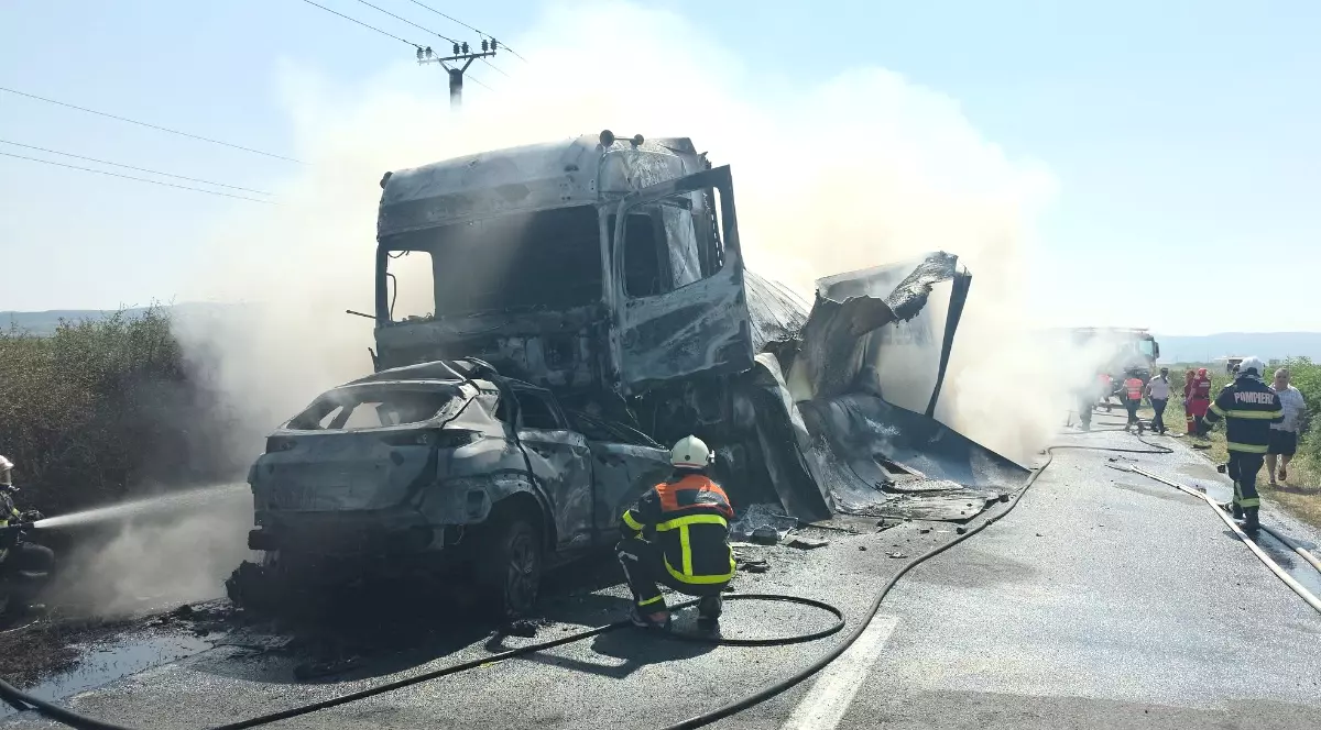 Imaginea prezintă pompierii intervenind la locul unui accident rutier grav, unde un autoturism și un camion au fost complet distruse de un incendiu puternic pe o șosea.