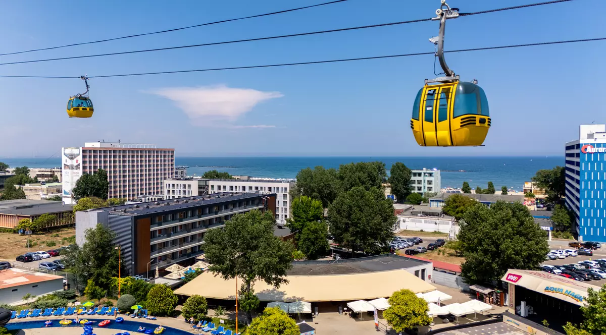 perspectivă aeriană asupra stațiunii mamaia, județul constanța. În partea superioară a cadrului se întind mai multe cabluri de suspensie pe care glisează două cabine de telegondolă cu ferestre largi, una fiind surprinsă în prim-plan, în dreapta, iar cealaltă la distanță, în stânga. Dedesubt, peisajul urban este dens, compus din diverse clădiri masive cu multe etaje, cu fațade geometrice și șiruri de balcoane. Construcțiile sunt intercalate cu porțiuni de vegetație deasă, cu copaci înalți, o parcare plină de autoturisme și o clădire joasă cu o copertină amplă, ușor curbată. În colțul din stânga jos se distinge marginea sinuoasă a unei piscine înconjurate de zeci de șezlonguri, iar în dreapta jos se află intrarea marcată a unui parc acvatic. În planul îndepărtat, dincolo de linia complexelor hoteliere și a unei fâșii de plajă presărate cu umbrele, orizontul este dominat de o întindere vastă de apă.