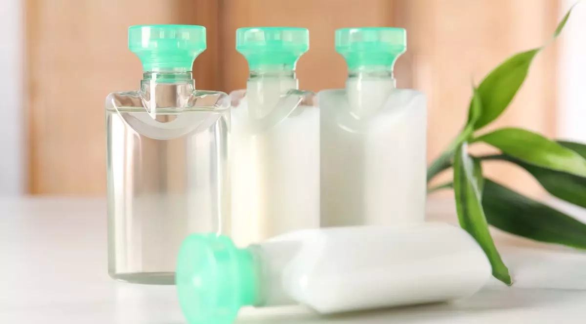 Sticle de plastic dintr-un hotel, cu produse cosmetice, transparente, și cu capace de culoare verde, pe fundal crem