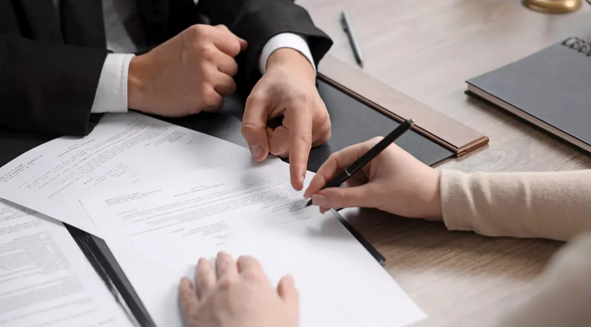 Imaginea surprinde momentul în care o persoană semnează un document în prezența unui profesionist, într-un cadru specific unei proceduri notariale sau juridice. Fotografia ilustrează procesul de încheiere a unor acte oficiale, fiind potrivită pentru subiecte legate de succesiuni, moșteniri sau alte formalități legale.