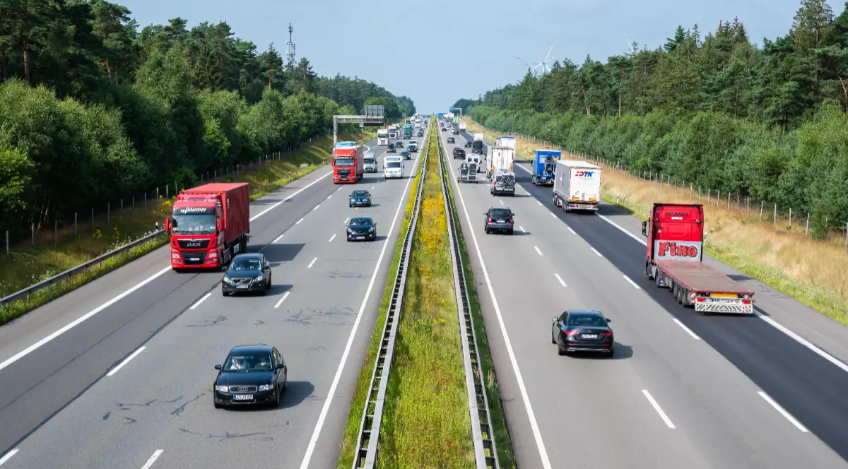 Imaginea prezintă o privire de sus asupra unei autostrăzi aglomerate, pe care circulă numeroase camioane și autoturisme în ambele sensuri, traversând o zonă împădurită.