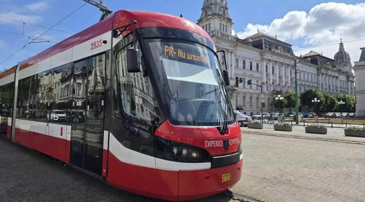 Tramvai de culoare roșie, în centrul municipiului Arad, cu cerul albastru pe fundal și o clădire istorică
