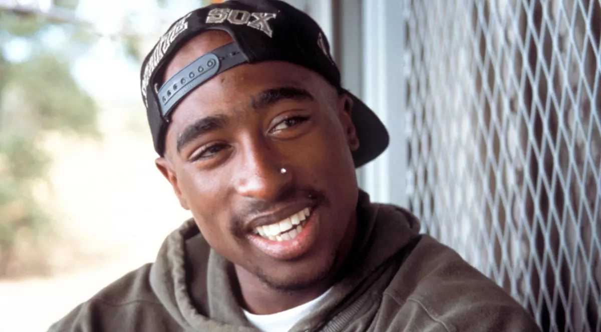 Imaginea prezintă un portret de aproape al celebrului rapper Tupac Shakur, zâmbind și purtând o șapcă orientată în spate și un hanorac verde.