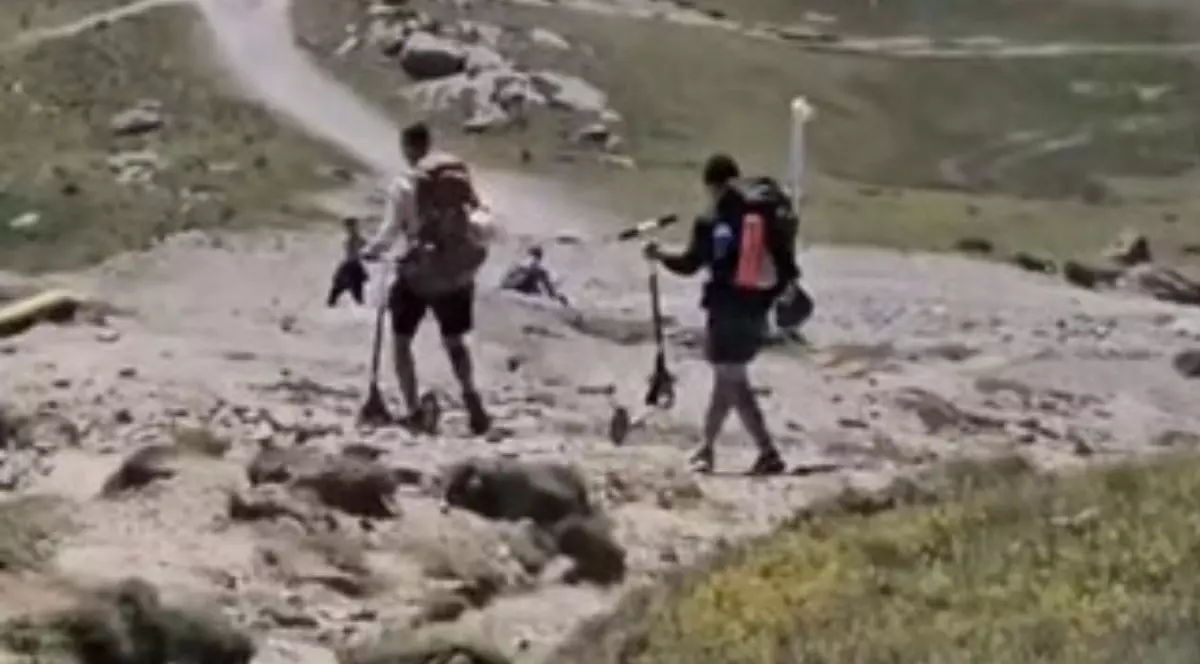 o scenă distribuită de Salvamont Prahova, în care apar turiști cu trotinete pe platoul Bucegi. Cadrul surprinde din spate două persoane care înaintează pe un teren montan accidentat, presărat cu pietre, bolovani și porțiuni de pământ denivelat. Ambii drumeți sunt echipați cu pantaloni scurți și poartă în spate rucsacuri de munte foarte voluminoase. Fiecare dintre ei ține și manevrează cu dificultate câte o trotinetă peste obstacolele de pe traseu. În planul îndepărtat, peisajul se extinde într-o zonă vastă cu vegetație măruntă, străbătută de o potecă lată, șerpuitoare, în jurul căreia se disting câteva stânci masive și siluetele altor câțiva turiști aflați în depărtare.