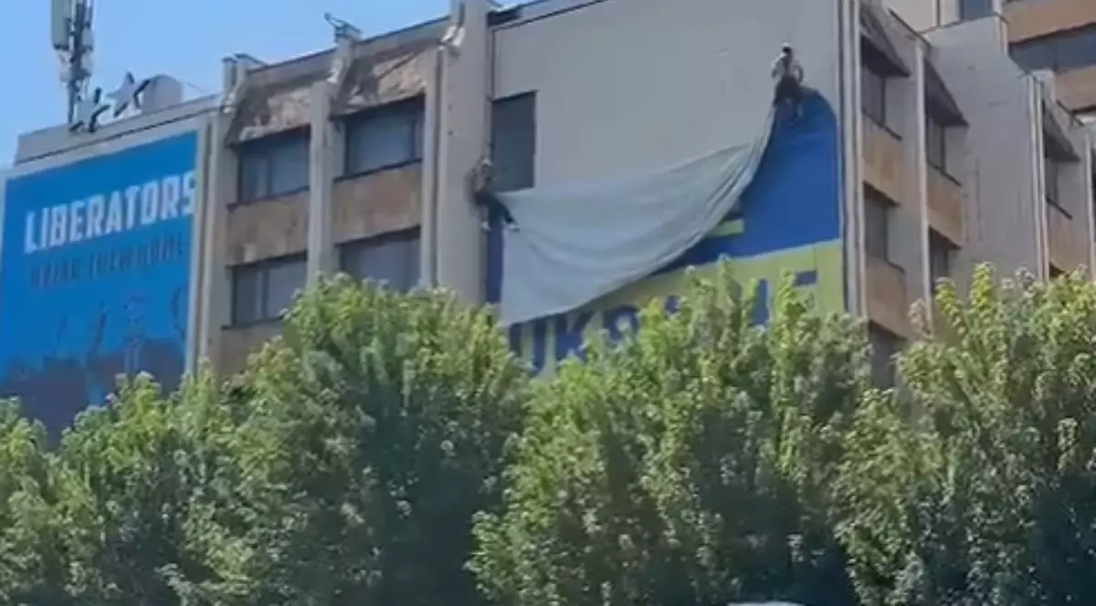 În imagine se vede o clădire din centrul orașului, pe fațada căreia este afișat un steag ucrainean de mari dimensiuni. Steagul este surprins în timp ce este coborât de pe clădire, în timp ce în partea laterală se poate observa un panou albastru cu mesajul „Liberators”.