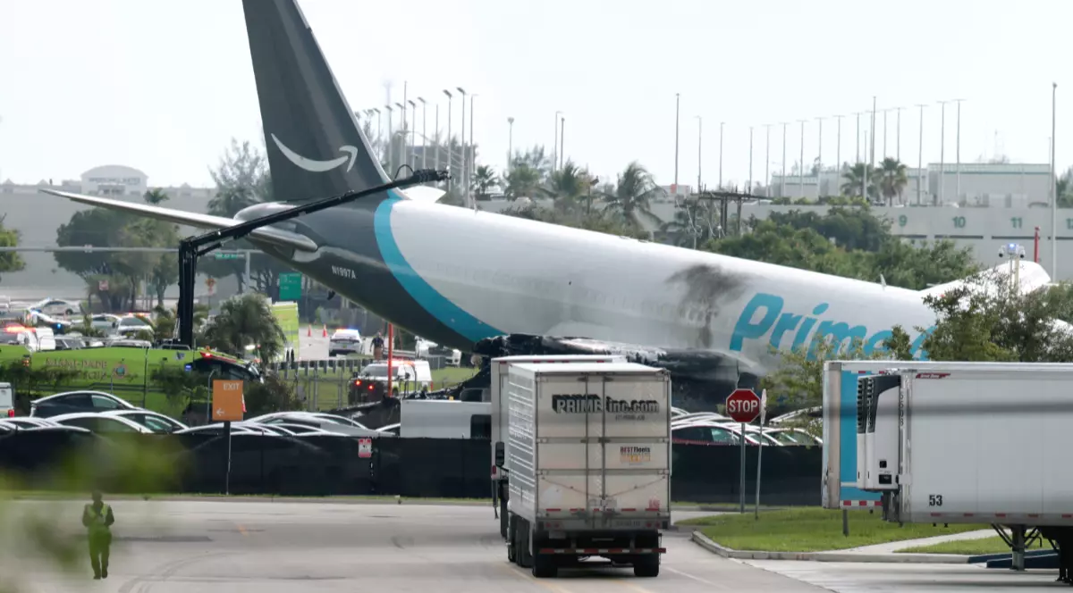 Imaginea prezintă un avion de marfă Prime Air avariat, oprit pe iarbă în apropierea unei zone de parcare, cu urme vizibile de arsuri pe fuzelaj.