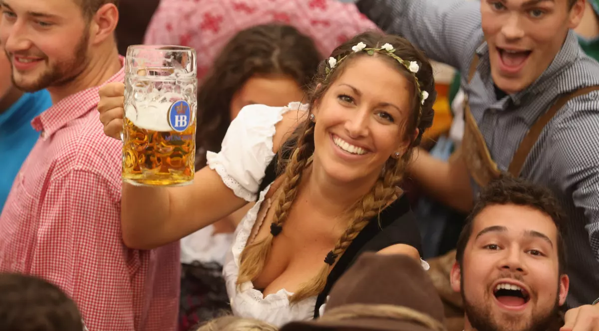 Mai mul'i tineri sărbătoresc la un festival în aer liber. În prim-plan, o tânără cu părul împletit împodobit cu o coroană de flori zâmbește senin, ținând în mâna stângă o halbă mare de bere.