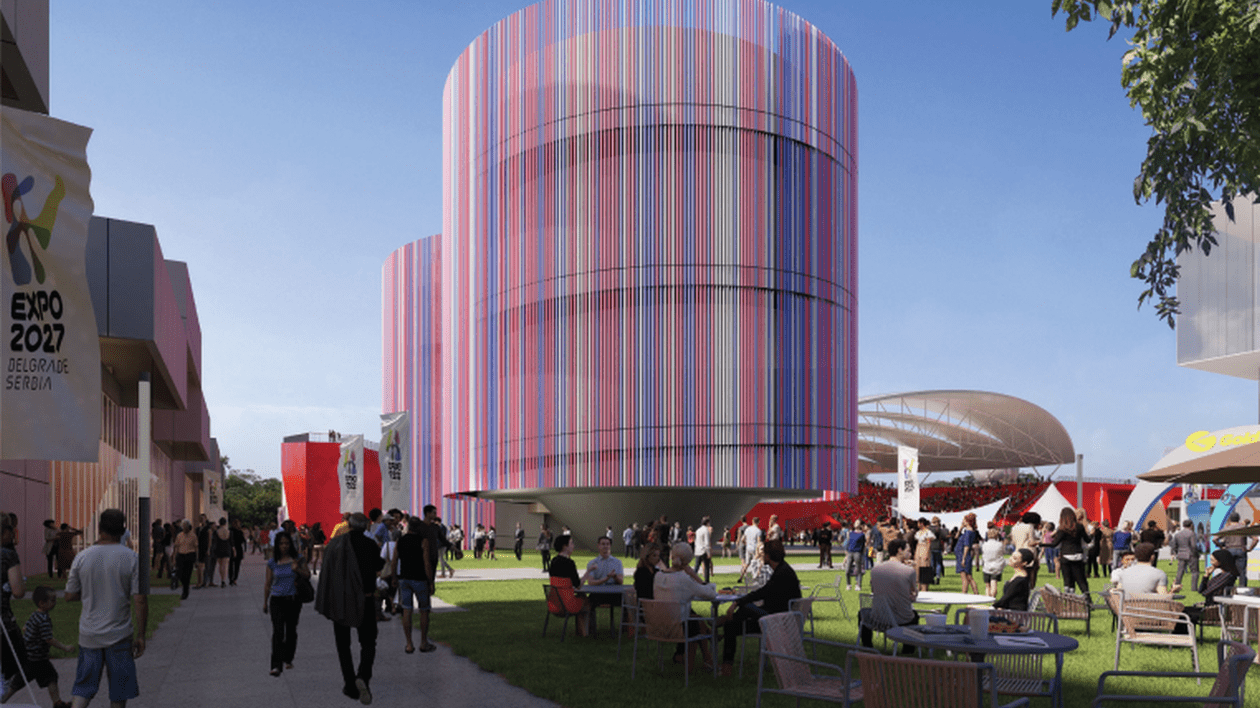 Mai mulți oameni se plimbă care se plimbă pe alei între spații verzi, la un centru expozițional, iar în stânga se vede pe un afișaj alb sigla Expo 2027