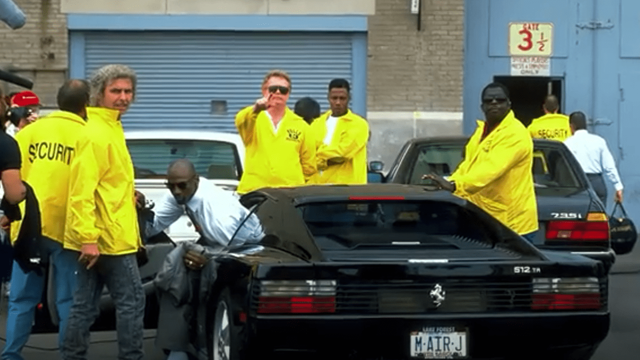 Michael Jordan coboară dintr-un Ferrari negru, înconjurat de agenți de securitate, îmbrăcați în geci galbene.