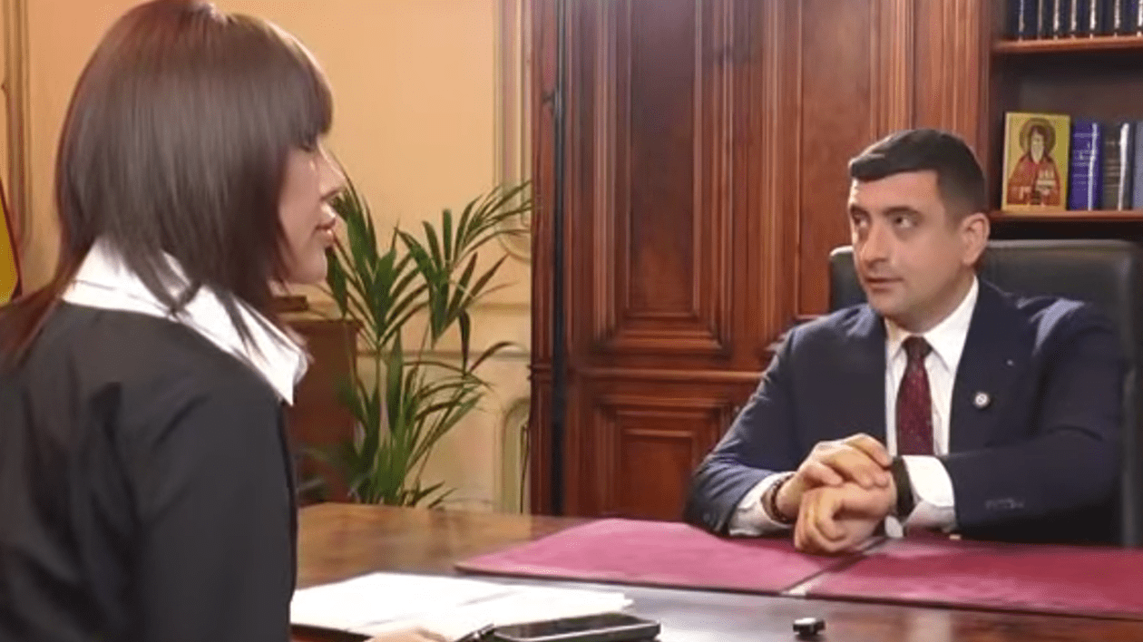 George Simion, șeezat la birou arătând spre ceasul de la mână. Deenise Rifai, pe scaun, cu spatel la cameră, discută cu el