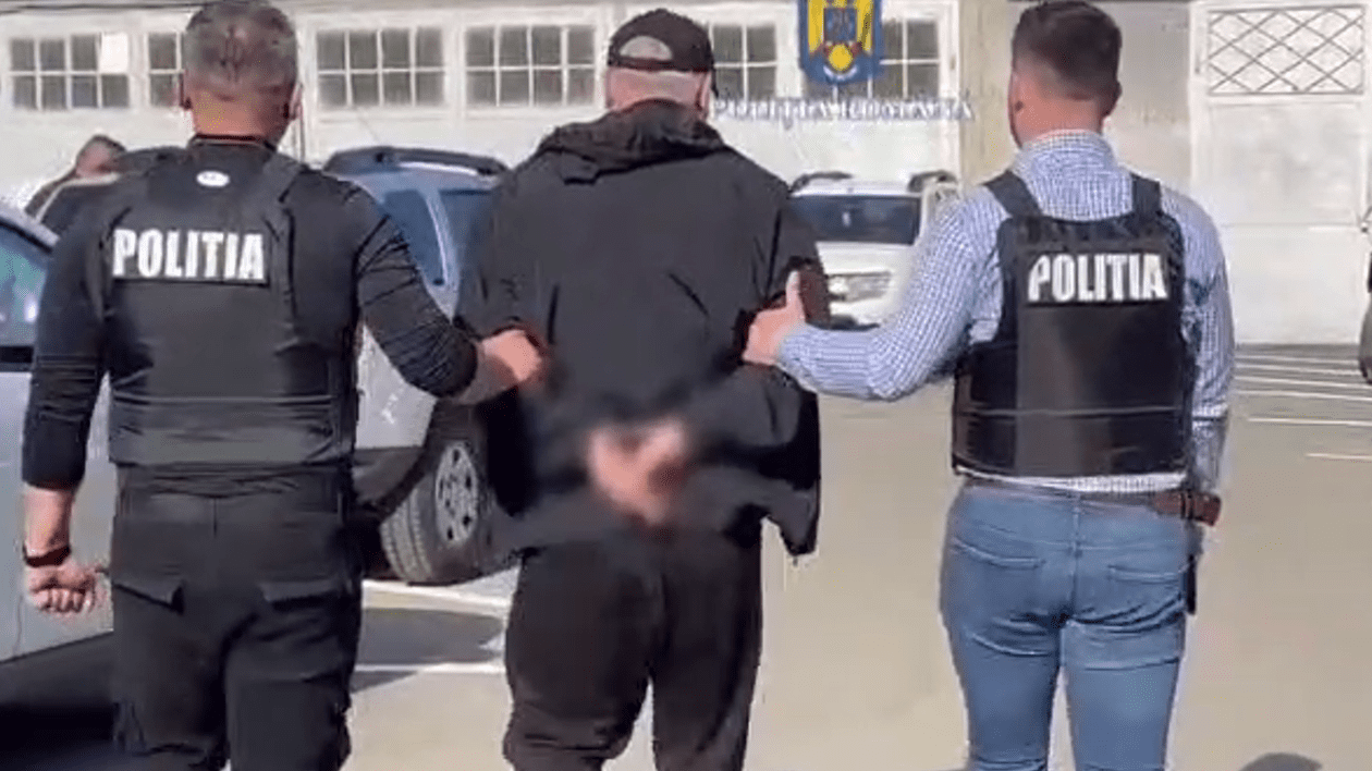 Un bărbat cu șapcă încătușat este flancat de doi polițiști. Toți sunt cu spatele și merge spre o clădire