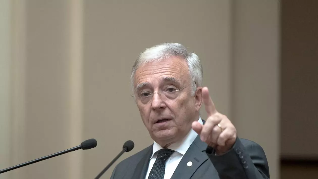 BNR prezintă raportul trimestrial asupra inflaţiei. Mugur Isărescu, la un pupitru, cu mâna stângă ridicată și degetul arătător în aer