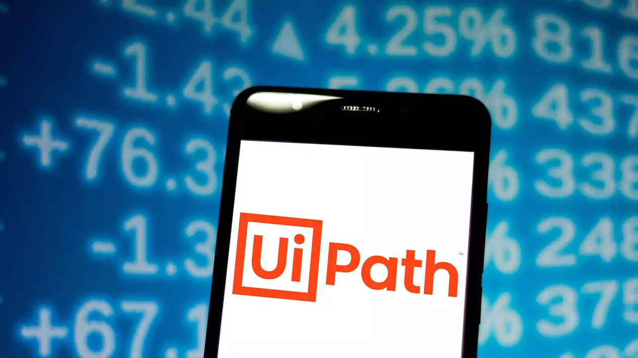 UiPath a strâns 1,34 miliarde de dolari pe bursă. Compania, evaluată la 29 de miliarde de dolari, mai mult decât Carrefour și aproape cât Panasonic. Logo-ul UiPath pe ecranul unui smartphone, cu cifre în spate