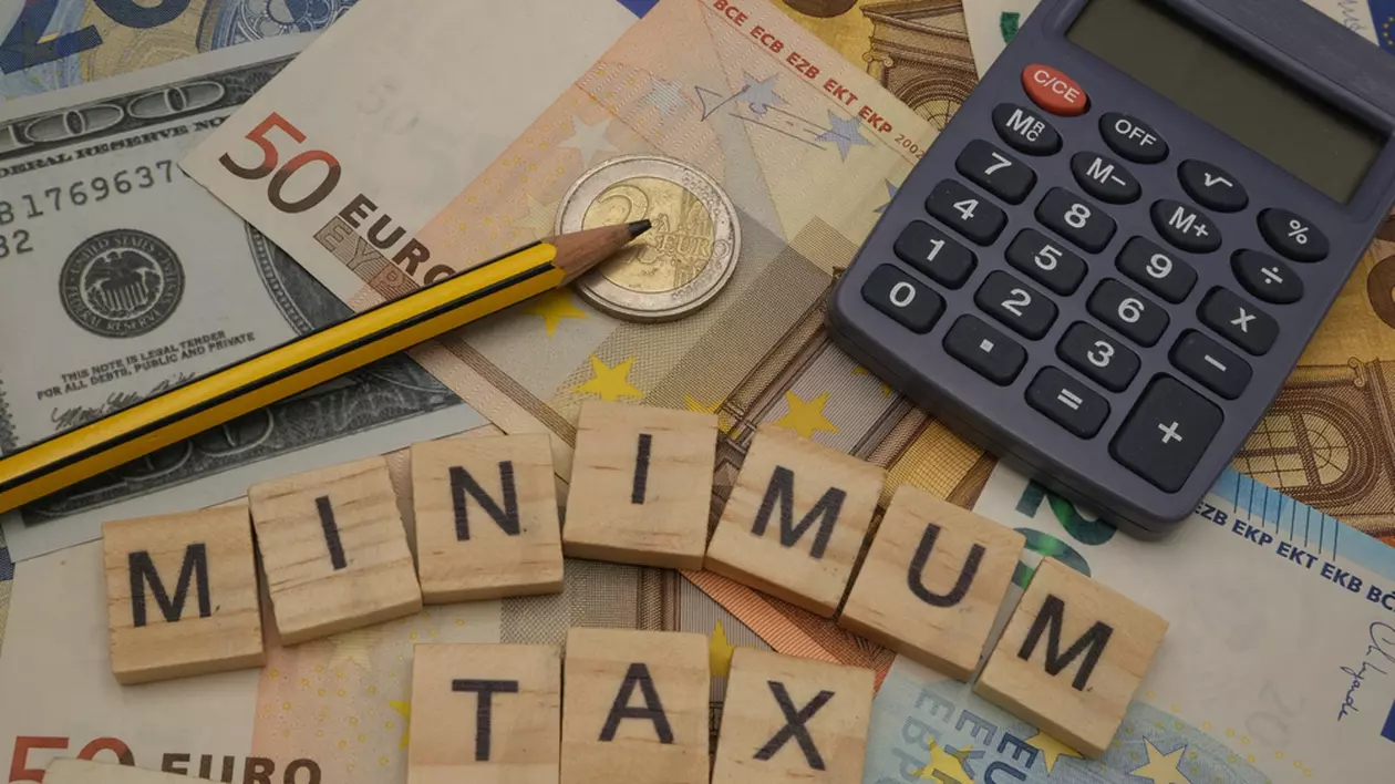 Care sunt ţările cu cele mai mici taxe din Europa - Imagine cu literele care formează cuvintele Minimum Tax alături de un calculator şi un creion pe un teanc de euro
