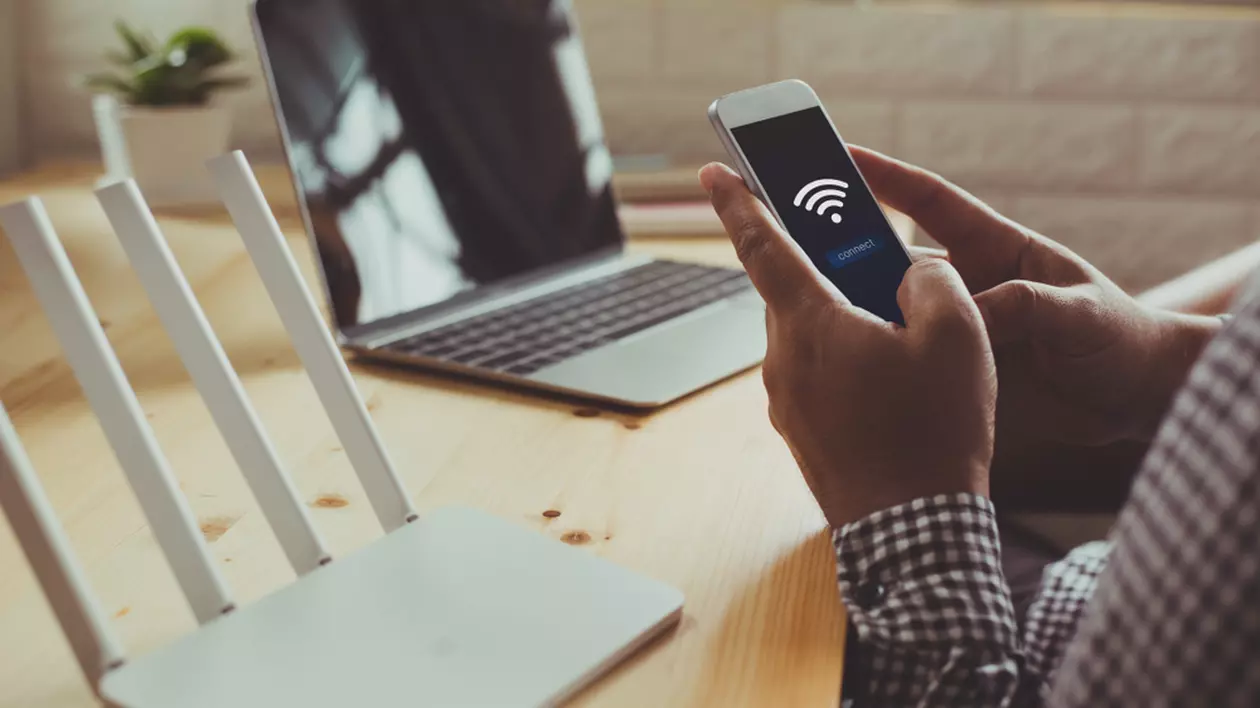 Cum alegi cel mai bun router Wi-Fi - Imagine cu un bărbat care ţine în mână un telefon mobil, având în faţa lui un birou pe care se află un laptop şi un router Wi-Fi