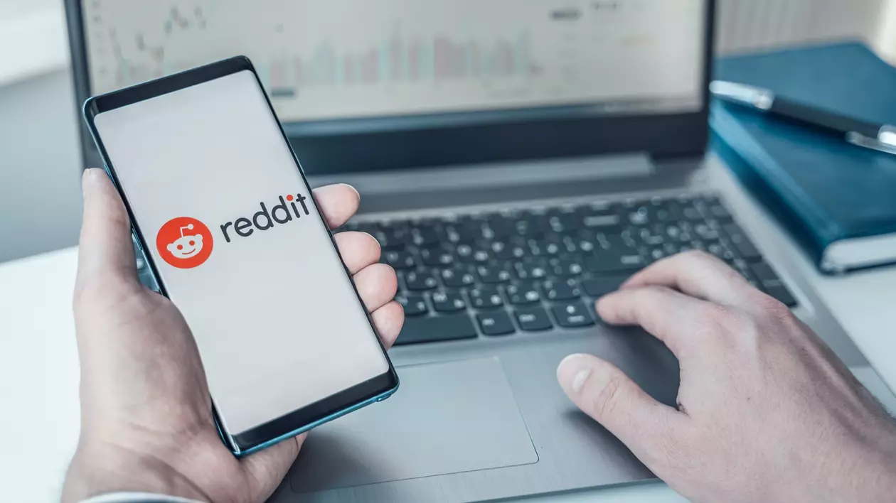 Ce este platforma Reddit şi cum să o foloseşti - Imagine cu un bărbat aflat în faţa unui laptop şi care ţine un telefon ce afişează logo-ul reddit