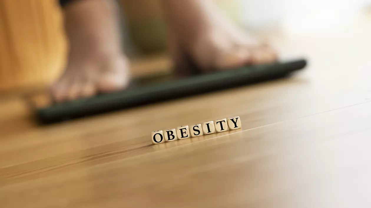 Imagine cu o persoană care se cântăreşte şi care are în faţă, pe podea, cuvântul obesity format din cuburi