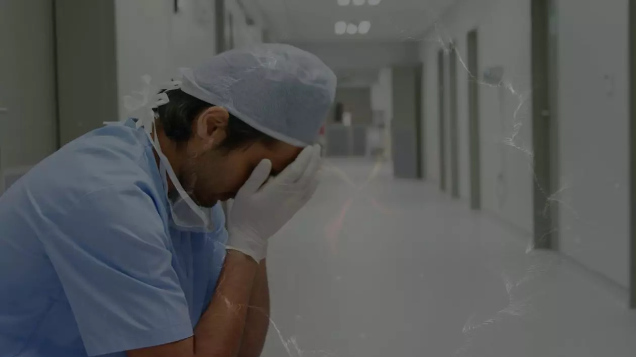 Imagine ilustrativă cu un asistent medical, îmbrăcat în uniforma de culoare albastră, cu o bonetă albă pe cap, ținând ambele mâini, pe care poartă mănuși, peste față