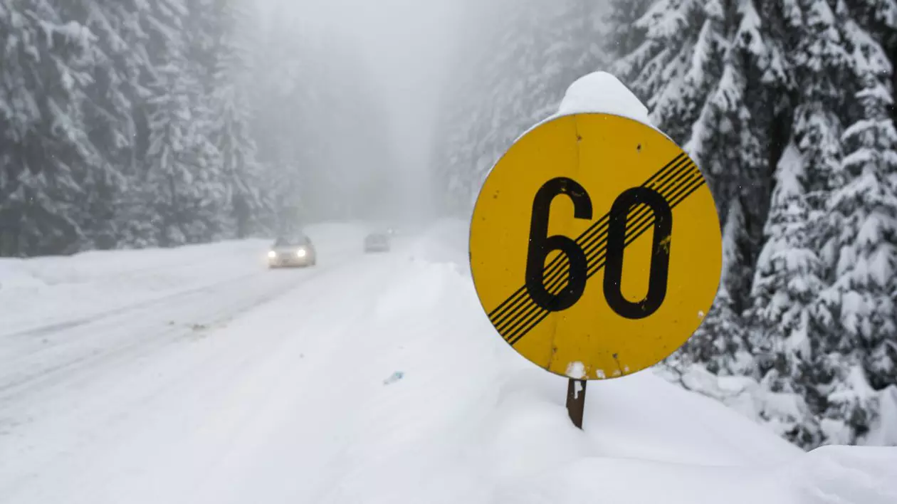 Drum de munte acoperit cu zăpadă și un indicator rutier de culoare galbenă de încetare a limitării vitezei la 60 de kilometri pe oră
