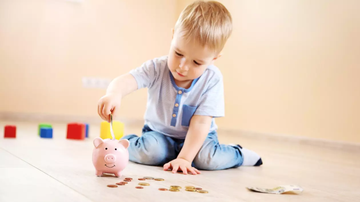 În imagine este ilustrat un copil care stă pe podea și pune bani în pușculiță