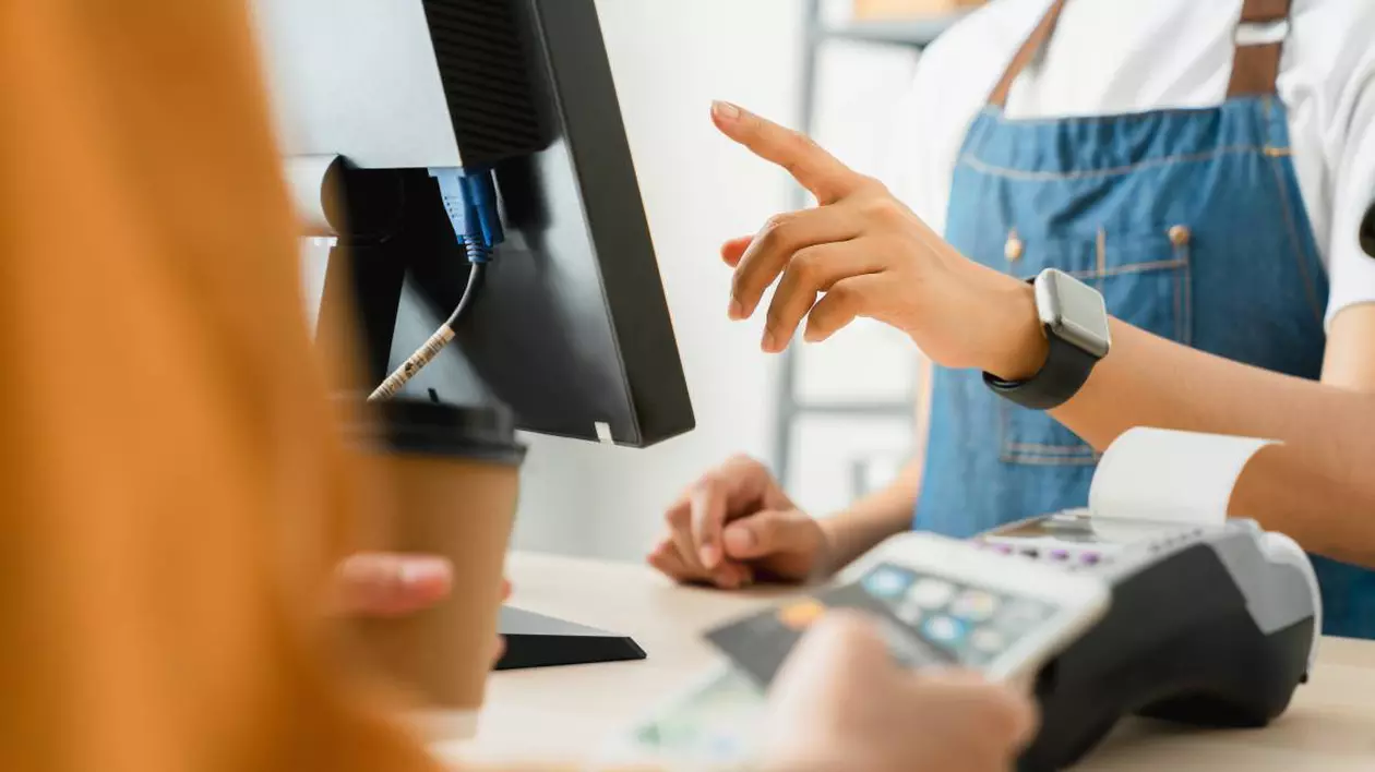 angajat al unui magazin care poarta un sort albastru tasteaza pe un monitor al unui computer. pe masa se afla un aparat pentru plata cu cardul