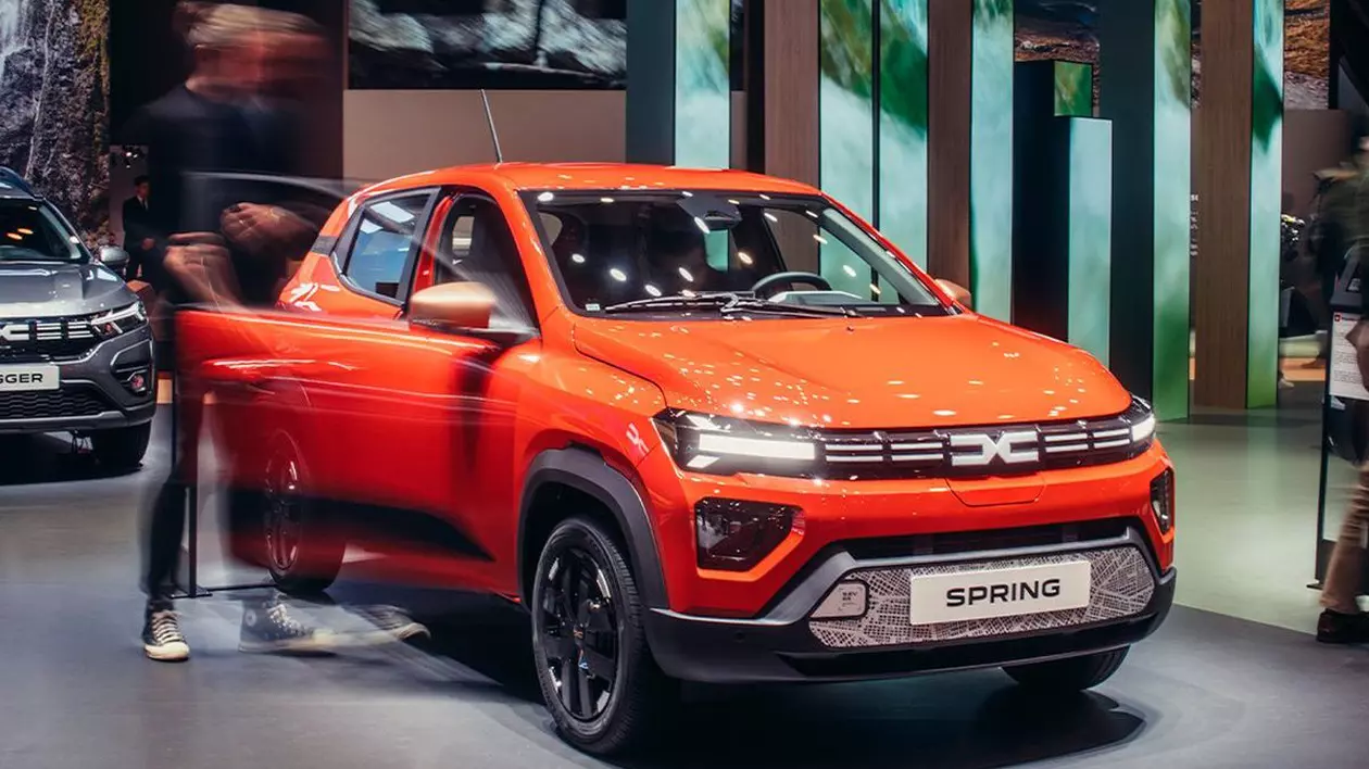 O mașină Dacia Spring portocalie, expusă la un salon auto, are o ușă deschisă, lângă care se află un bărbat