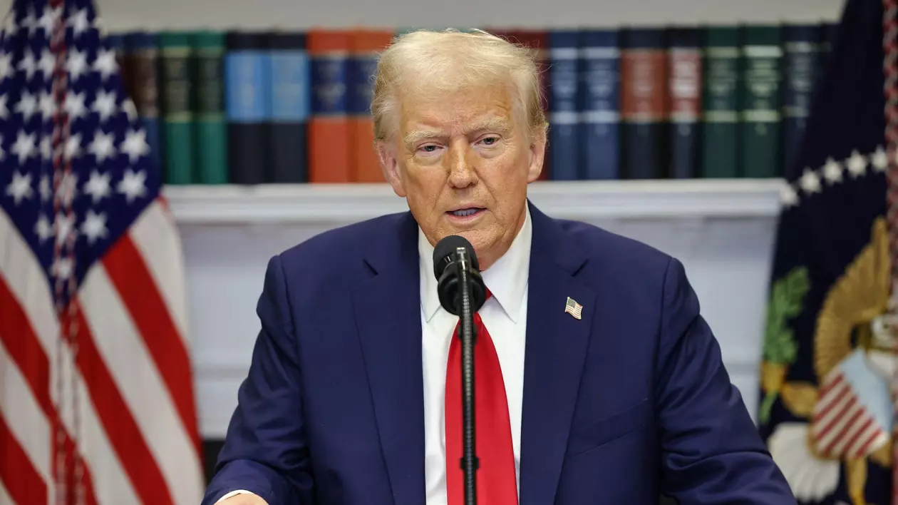 Donald Trump, într-o conferință de presă la Casa Albă, îmbrăcat într-un costum bleumarin cu o cravată roșie, așezat în fața steagului Americii