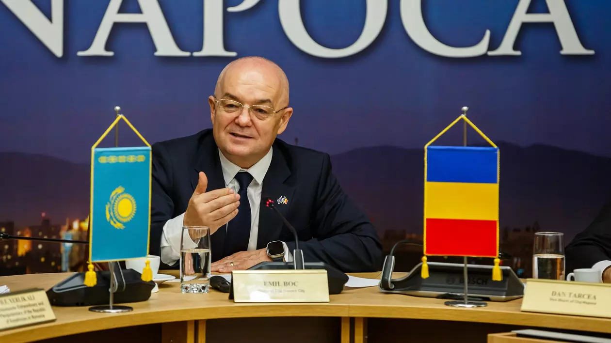 Primarul din Cluj, Emil Boc, stă la o masă într-o sală în care are loc o conferință de presă, iar lângă el este steagul României