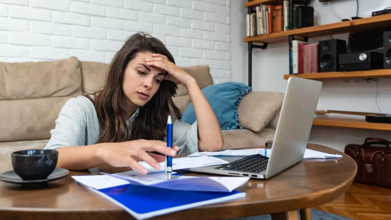 O femeie stresată, cu mâna la cap, se uită la niște facturi așezate lângă laptop, pe o masă