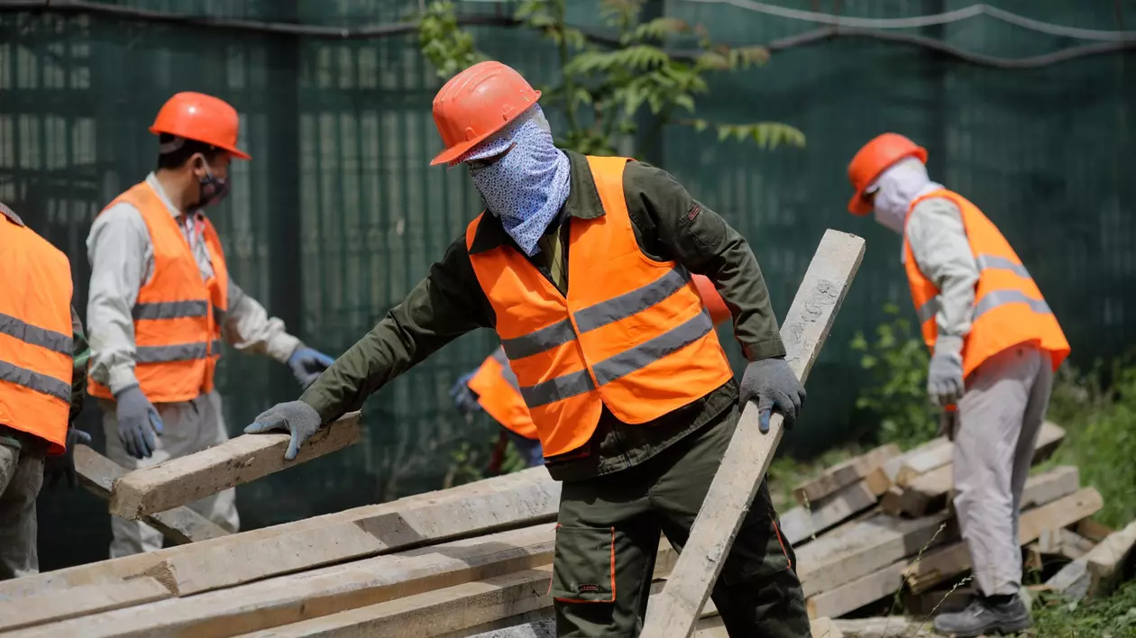mai mulți muncitori străini, cu veste portocalii și căști de protecție lucrează în construcții