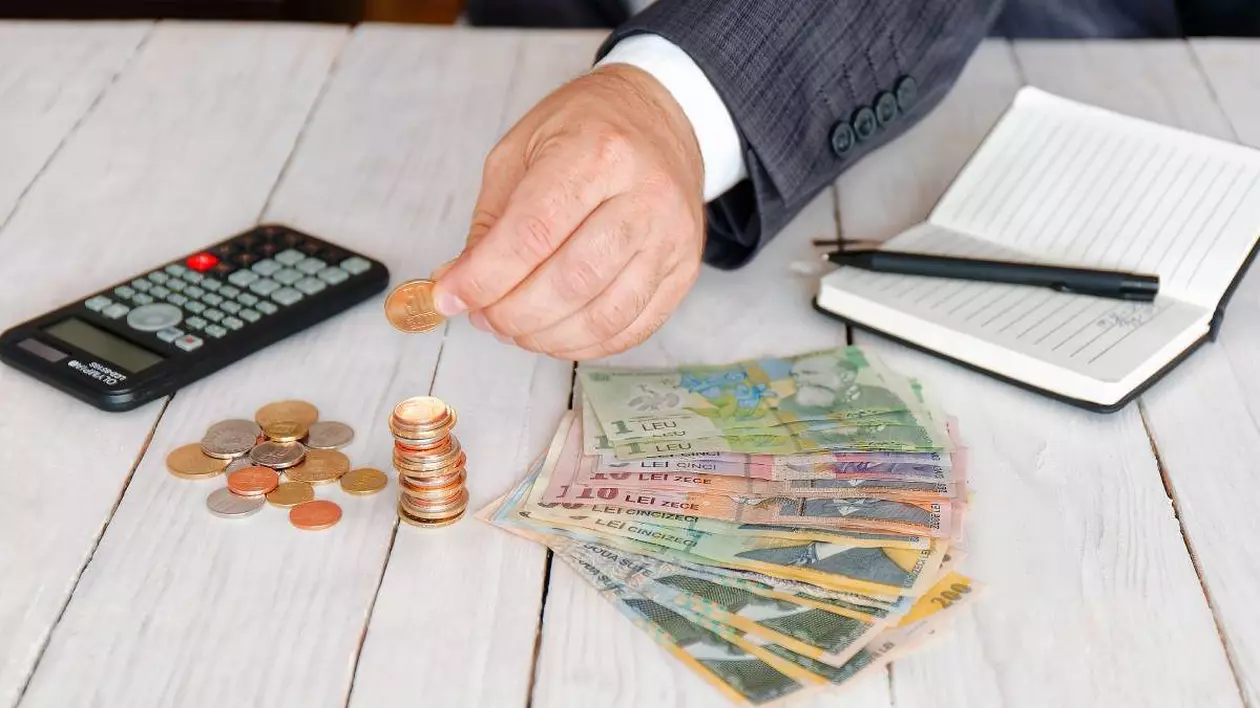 Mâna unui bărbat care pune o monedă într-un teanc de bani, pe o masă pe care mai sunt bancnote, un calculator și o agendă cu pix