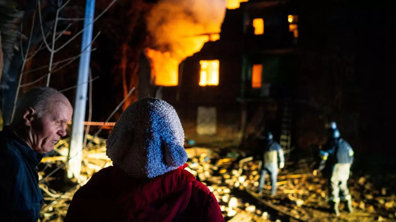 O persoană cu spatele, care poartă o cârpă pe cap, se uită la doi pompieri care încearcă să stingă flăcările la o clădire bombardată în Ucraina, pe timp de noapte